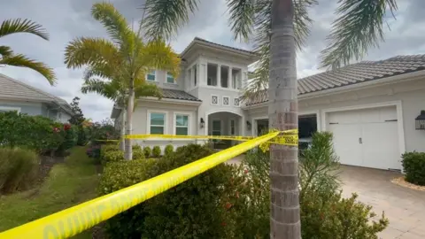 Manatee County Sheriff’s Office A cream Florida home, with a number of palm trees in the foreground. There is yellow police tape wrapped around the trees. In the garden in front of the house there are a range of bushes, and a patio car parking space.