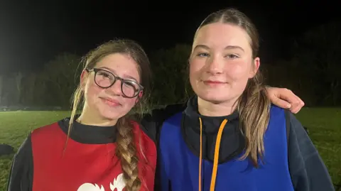 ELLEN KNIGHT/BBC Esme and Megan stood side by side, smiling slightly and looking into the camera. They're both wearing black sweatshirts - Esme, on the left, is wearing a red tabard, and Megan, on the right, is wearing a blue one. They're stood on a flood-lit rugby pitch, which has green grass, a dark night sky, and is bordered by trees.