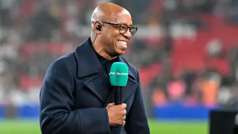 Getty Images Ian Wright stands pitchside holding an ITV Sport-branded microphone. He's wearing a heavy coat and smiling at someone off-screen.