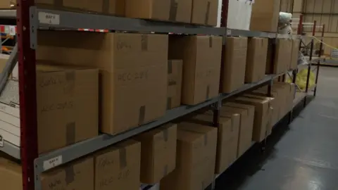 Linsey Smith/BBC Brown cardboard boxes are stacked on to metal shelves in a warehouse in Reading, Berkshire.