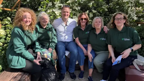 Gary Morrisroe David Maxwell, sitting in a garden surrounded by five women from the Strabane Community Project. He is smiling and wearing blue jeans and a open-necked white shirt. The women are all wearing dark green polo shirts with a "Strabane Foodbank" logo