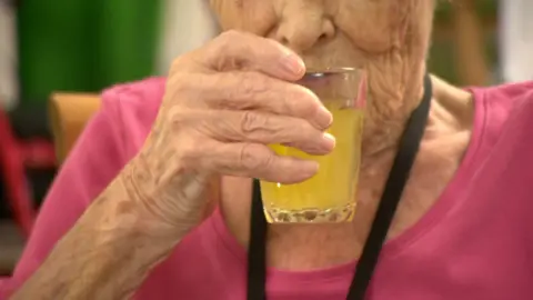 An elderly woman dressed in pink sips from a glass of orange liquid.