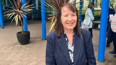 BBC/RICHARD EDWARDS Angela Rushton, who spoke against the 1,500 home plan at the meeting. It shows her standing outside North Yorkshire Council's offices in Selby. She is looking slightly to the right of the camera, has brown hair and is wearing a blue blazer. A big, yucca-type plant is just behind her in the shot.