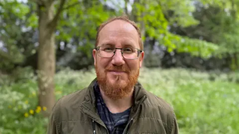 Damian is in the centre of the photo. He has ginger hair and a ginger beard, and is wearing glasses. He's wearing a green jacket, and a blue checked shirt. In the background to his left is a tree, among a meadow of wild flowers.