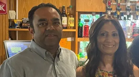 FLAME RESTAURANT/WILBURTON Two people, standing side-by-side behind a pub bar. Dinesh Makwana, on the left, is a man with dark hair swept back. He is wearing a khaki shirt buttoned up to the neck. On the right, his wife, Harsha, has long, brown shoulder-length hair and a floral design dress. Bottles can be seen behind them on a shelf. Pub optics are also visible.