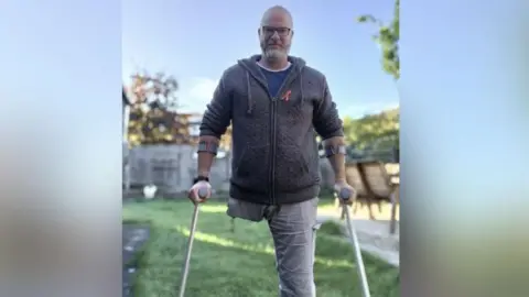 Picture of Pete Lloyd in garden. He has one leg (left leg) and is on crutches. He has a dark blue jumper on and light jeans.