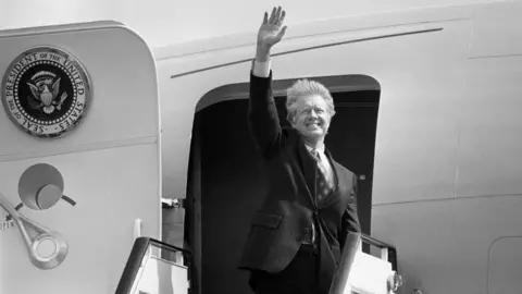 Jimmy Carter waving at the door of Air Force One which has the president's seal on the door - a circular badge with an eagle in the centre. Carter has grey, windswept hair.