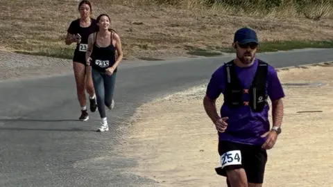 A man at the front of the picture is running, wearing a blue t shirt and a black weighted vest. He is wearing black shorts and has the number 254 on his shorts. Two women are in the back of the picture, runing and smiling wearing black tops and one is wearing grey leggings, with one in black shorts. The ground behind them all is very dry grass.