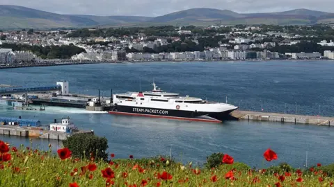 MANX SCENES A view from Douglas Head of the Manannan ferry moored in Douglas Harbour. There are hills and the sweep of Douglas Bay with its Victorian Promenade in the background, and poppies on a grassy bank in the fore.