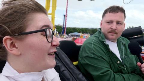 A woman with glasses is on a rollercoaster with a male reporter. He holds a mic, wears a green shirt and has short dark hair and a moustache