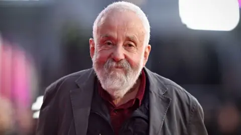 Getty Images Mike Leigh looks at the camera. He has a white beard and mustache and is wearing a gray jacket and two layers underneath