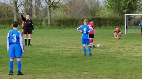 Ernie Broughton signals for players to wait as a defender, near a goal, laces up his boots