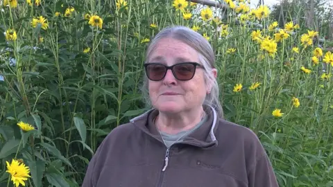 Jenny Morgan – wearing a brown fleece and sunglasses. Behind her are wildflowers on a grassy bank.