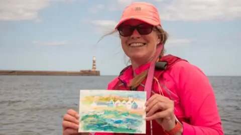 Anita Jazmin holds her painting to the camera, with the sea and a lighthouse behind her
