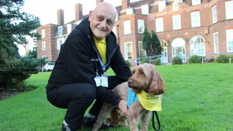 NHS Humber Health Partnership Denis is crouching down holding Banrey underneath his tummy on a green lawn in front of a large, red-brick hospital building.  Barney wears a yellow neckerchief which identifies him as a therapy dog.