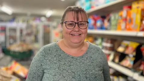 Martin Giles/BBC Julie Woods has dark hair in a ponytail and brown-rimmed glasses. She is wearing a green patterned top and is standing in front of food shelves.