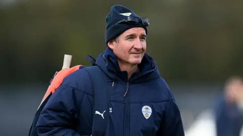 Mal Loye wears a navy woolly hat, puffer jacket and carries an orange bag with a cricket bat sticking out of it. He has sunglasses on his head. 