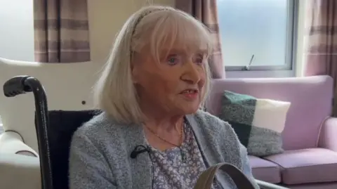Ruby Lee wears a blue cardigan and is looking off camera. The room behind her has a purple sofa and purple patterned curtains