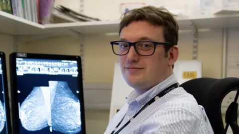 CUH/NWAFT Dr James Tanner is off-centre to the right. He is sitting on a black chair at computers which are showing a breast screening image. He has short brown hair and is wearing black framed glasses, a blue striped shirt and a black lanyard.