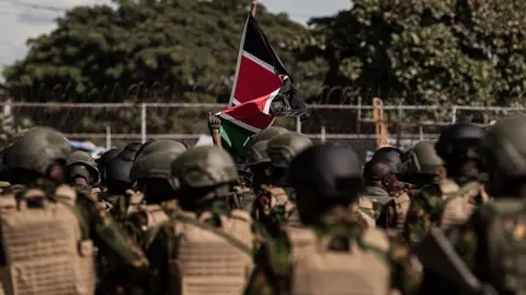 A Kenyan flag unfurls above a group of people in khaki police uniforms with their back to the camera. They are out of focus.