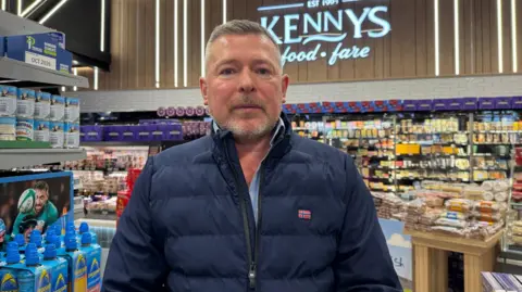 Kenny Bradley stands in his supermarket, behind him in the far distance fridges with cheese in them can be seen, to his right are bottles of sports drinks and also canned goods. Kenny has short grey hair and a short grey beard, he is wearing a dark blue padded jacket with a small Norwegian flag on its left breast.
