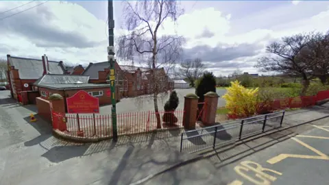 Google The former site of Trimdon Grange Infant and Nursery School, viewed from the main road. In the foreground is fresh red painted gates and rails, with shrubbery. The playground is in front of the single-tier school building and a small grassed play area is on the right. It is a sunny spring day with wispy clouds overhead.