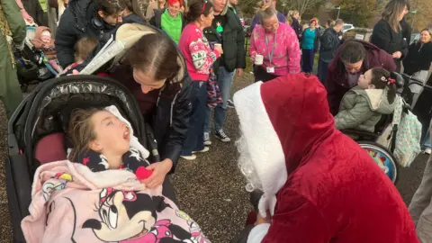 A girl in a wheelchair covered in a blanket smiling at Father Christmas