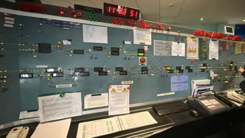 Network Rail An old train signalling system which has a clock at the time. There are lots of buttons and panels, and there are books on the table.