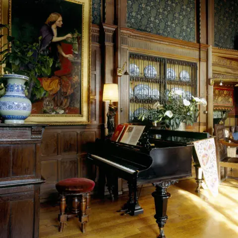 National Trust Images/George Frederick Watts A room with a black grand piano and an oil painting on the wall. The painting shows a woman with long blonde hair and a blue dress.