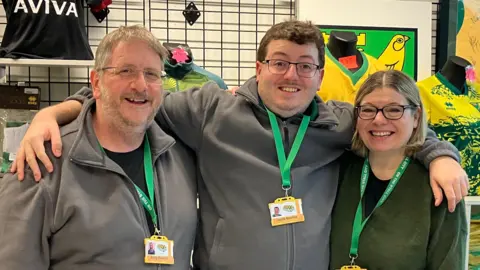 Members from the On The Stall City shop standing in a row looking directly at the camera and smiling. Andy Bowles is on the left dressed in a black T-shirt and grey zip up sweatshirt. He is wearing a pair of glasses and has a green lanyard round his neck. 