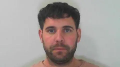 North Yorkshire Police A man, in his forties, wearing a salmon-coloured top, with black hair and a short beard looks directly into the camera, stood in front of a white backdrop.