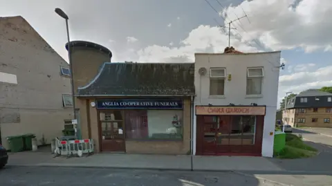 Image from 2014 showing Anglia Co-Operative Funerals shop next to a China Garden takeaway. There is some building work on the left corner of the building with a temporary fence in place