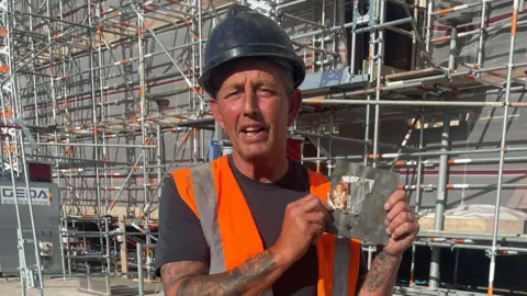 Alan Needham wearing a hard hat and orange high-visibility vest stands in front of metal scaffolding, holding up a square slate with a damaged photo attached.