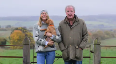 A woman with long blonde/brown hair and wearing a grey jumper is next to a man in a coat. They are both wearing blue jeans and standing by a gate in a field.