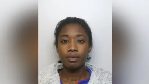 A woman in her twenties pictured in a police mugshot. She is wearing a grey fluffy jacket and a blue top underneath. She is staring directly at the camera.