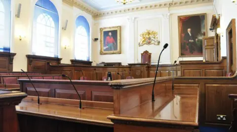 BBC Guernsey States Chamber. The room has cream coloured walls with large paintings encased in gold frames. The room is filled with dark wood where people can sit with large black microphones fitted to each table.