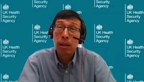 Prof Andrew Lee wearing a headset, with dark hair and a blue shirt on, in front of a UKHSA blue background