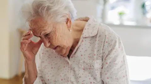 Getty Images An elderly woman in white pyjamas with a pink floral design sits on her bed, the background is out of focus, and she holds her hand to her head as if she's experiencing a headache or has forgotten something