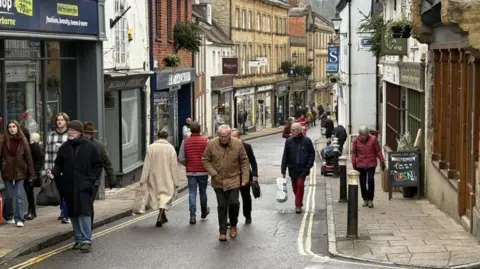 Shoppers walking up and down Cheap Street in Sherborne. It is winter and it has been raining so the road and pavement are wet. People are walking on the pavement as well as on the street. There are shops on either side of the road.