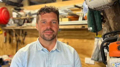 Joel Freire is standing in his tree surgery shed, with a wooden workbench of tools behind him and the edge of a chainsaw visible to the right of the frame. He is wearing a pale blue shirt and looking into the camera.