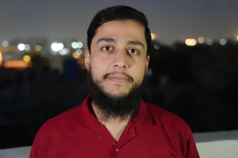 Sh Ed Kamil Khan Abu Bakar Ahmad smiles looking directly at the camera, he has short black hair and a black beard. He is wearing a red shirt with the collar open at the top. He is pictured on what appears to be a rooftop, with the night's sky in the background.