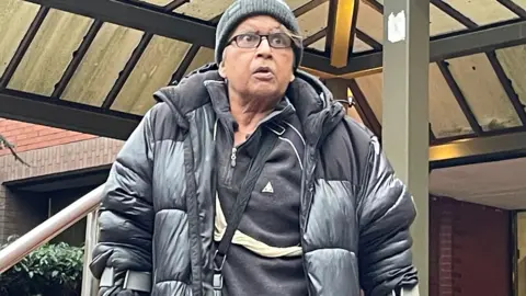 PA Media Farhat Ajaz, 62, leaving Birmingham Crown Court. He is wearing a black puffer jacket and woolly hat and is wearing glasses and walking with crutches.