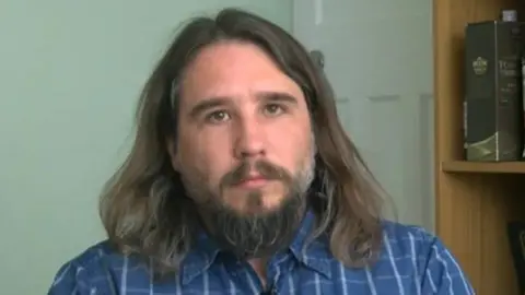 BBC Robert Miller wearing a blue chequered shirt with a collar. He is a white man who has long brown hair and a long dark beard.