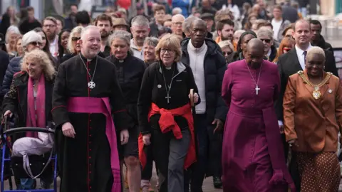 The Archbishop of Canterbury walking towards the camera with other religious leaders and 100 people behind her