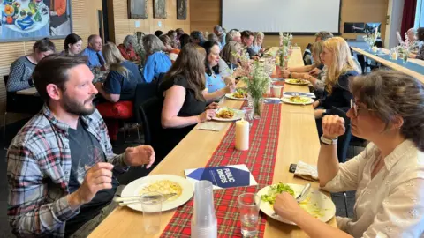 A group of people sitting at a long dinner table. The guests are sitting across from each other in a well lit room. The table features a red tartan centrepiece with false candles and flowers on it.