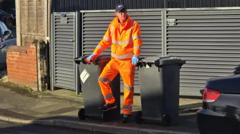 Hayley Coyle/BBC Bin loader Rick Brown stood on a street with both hands on two grey bins waiting for the bin lorry 