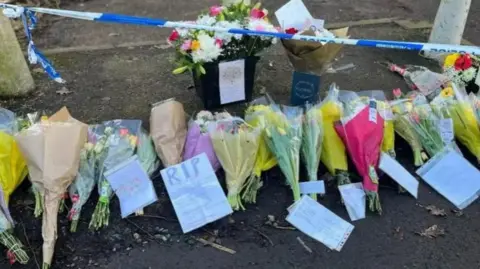 Floral tributes lie in a line, at the scene of Leo's stabbing. There is blue and white police tape at the top of the shot