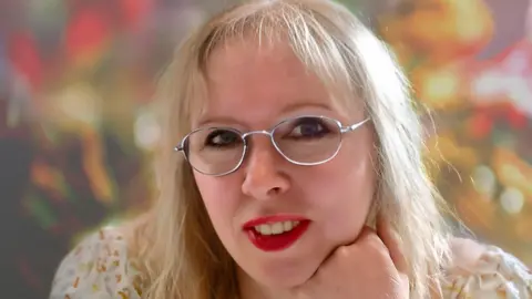 Michele Romano smiling at the camera and wearing red lipstick. 
She is wearing glasses with a white frame