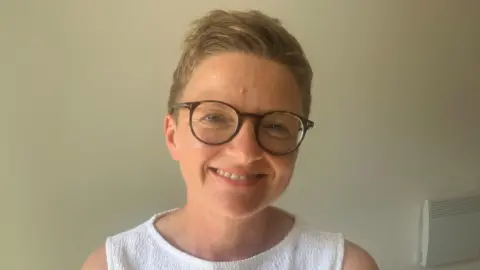 Mayor Bev Craig is smiling. She is wearing glasses and wearing a white vest top. There is a plain white wall in the background.
