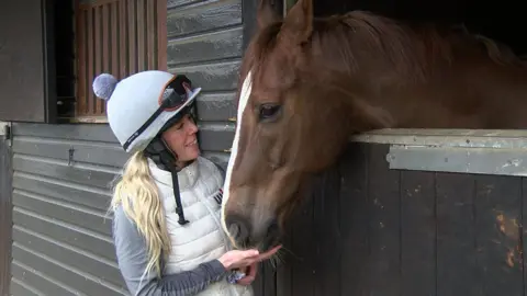 A women wearing a jockey uniform pets a brown racehorse who is looking out from a stable.